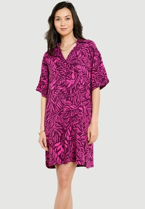 Žena nosi voľné krátke šaty po kolená s jasne ružovými a tmavofialovými vzormi listov na jednofarebnom pozadí.