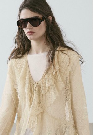 Donna con capelli castano lunghi che indossa grandi occhiali da sole tartarugati e una blusa beige con volant in pizzo sopra una maglietta bianca.