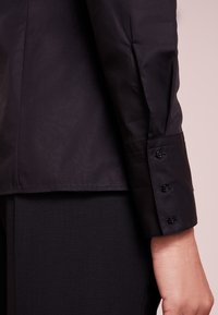 Camicia nera a maniche lunghe con polsini abbottonati e una texture liscia, con un taglio elegante e una leggera lucentezza su un tessuto nero a contrasto.
