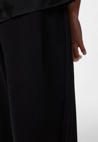 Črne hlače širokih nog z mehkim materialom, z gladko teksturo in stranskimi šivi. Na tkanini je vidna roka, ki dodaja kontekst.