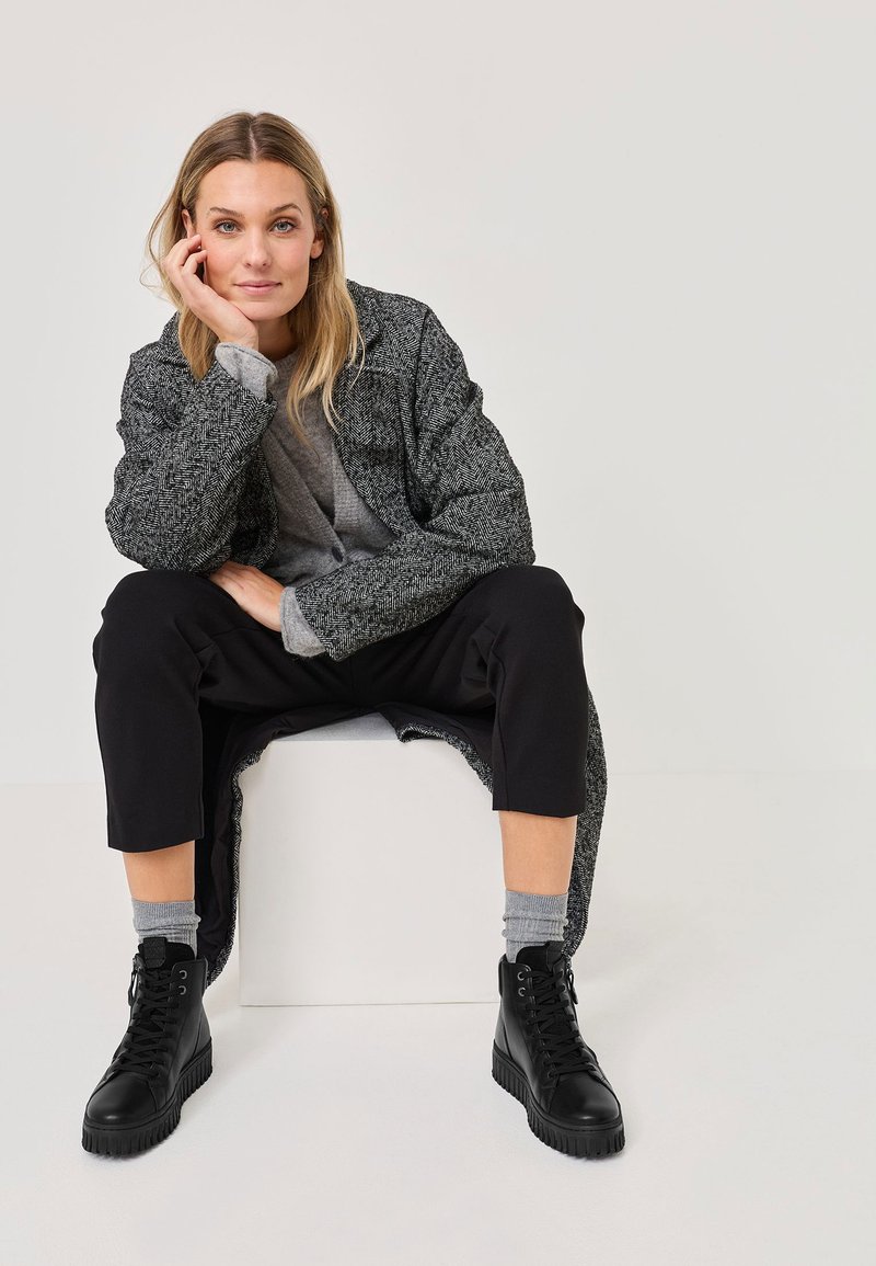 Dugi kaput s uzorkom hounds-tooth u sivoj boji preko sive majice, crnih krojenih hlača i crnih čizama na vezanje. Svjetlosive čarape su vidljive. Neutralna pozadina.