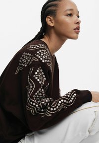 Hanorac maro cu modele complicate de broderie albă și bej pe mâneci, având un design relaxat și un material texturat.