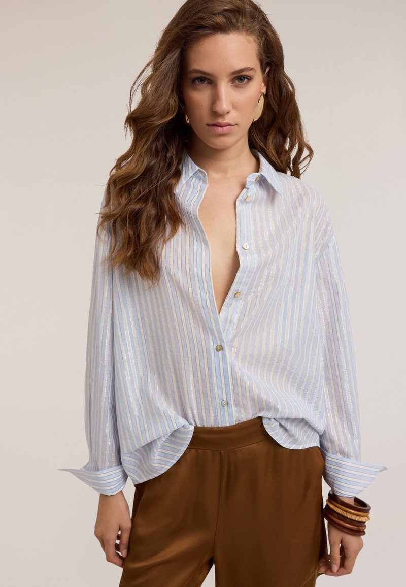 Camicia a righe blu chiaro con una vestibilità comoda, abbottonatura sul davanti e maniche arrotolate, abbinata a pantaloni in satin marroni e braccialetti sovrapposti.