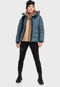 Puffige tealfarbene Jacke mit diagonalen Steppnähten, beigem Fleece-Layer, schwarzen Cargo-Hosen und schwarzen Schnürstiefeln. Akzentuiert mit einer grünen Strickmütze.