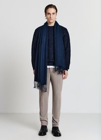 Sciarpa di lana blu navy con bordi sfrangiati posata su un maglione scuro, pantaloni beige e scarpe nere, esposta su uno sfondo neutro.