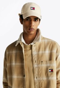 Camisa de cuadros en tonos beige y marrón con cuello de borreguillo, sobre un suéter beige, combinada con una gorra beige que lleva un logo.