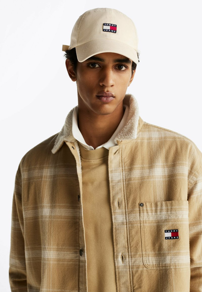 Camisa de cuadros en tonos beige y marrón con cuello de borreguillo, sobre un suéter beige, combinada con una gorra beige que lleva un logo.