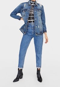 Denimjack met versleten details, blauwe kleur, vier voorzakken, gedragen met een zwart-wit gestreept topje en een high-waisted spijkerbroek.