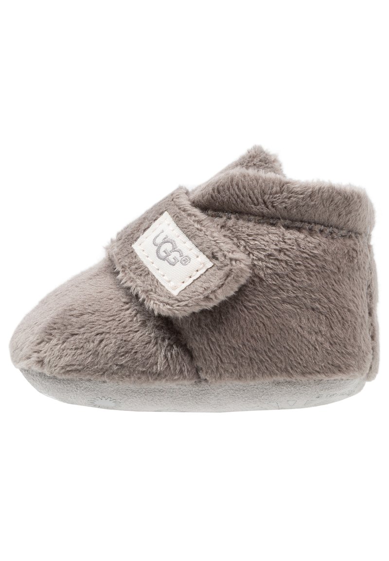Zapatilla gris para Niños, hecha de suave felpa texturizada, con forma redondeada y una correa de Velcro. Presenta una etiqueta de marca en el costado.