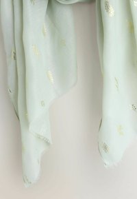 Bufanda verde claro con patrones de hojas doradas, hecha de tela suave y ligera. Los bordes tienen un fleco delicado para dar textura.