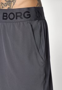 Nærbilde av grå elastiske bukser med "BORG" skrift på linningen og en sidelomme, båret av en tatovert person.