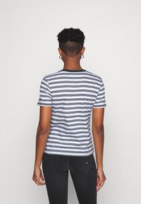 Person som bär en kortärmad vit och marinblå randig skjorta samt svarta jeans, stående vänd mot en helt vit vägg.