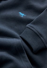 Mørk marineblå sweatshirtstof med ribbet manchet og lille broderet lyseblåt T-Rex dinosaurlogo på brystet.