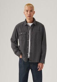 Chemise en denim gris avec deux poches avant, design à boutons et col. Portée sur une chemise blanche avec un jean bleu foncé. Texture lisse.
