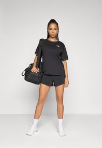 Kvinna i svart Puma träningskläder med shorts och t-shirt, bärandes en svart gymväska över axeln, stående mot en enfärgad bakgrund.