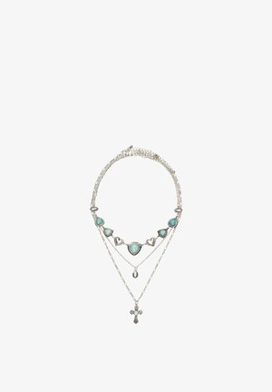 Collana a più strati in argento con pietre azzurre a forma di cuore, un piccolo pendente ovale e un pendente a croce dettagliato su sfondo bianco.