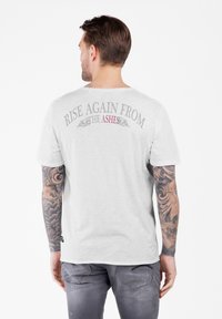 Mann mit tätowierten Armen, der ein weißes T-Shirt trägt, auf dem auf dem Rücken "RISE AGAIN FROM THE ASHES" gedruckt ist, kombiniert mit grauen Hosen.