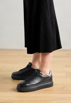 Jambes portant un pantalon large côtelé noir et des baskets plateforme en cuir noir, debout sur un sol en bois clair.
