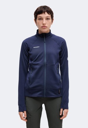 Ung voksen iført en mørkeblå lynlåstrøje med Mammut-logo, stående oprejst mod en ensfarvet lys baggrund.