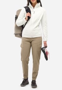 Personne portant un pantalon beige, une veste en polaire blanche, des chaussures de randonnée noires, portant un sac à dos beige et tenant une chaussure d'escalade rose et noire.