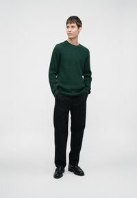 Groene gebreide trui met textuur en een ronde halslijn, gecombineerd met zwarte broek en zwarte leren schoenen. Eenvoudig, gestroomlijnd ontwerp.