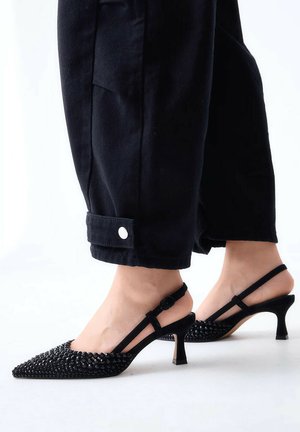 Piedi femminili che indossano décolleté slingback nere con punta appuntita e dettagli con borchie e pantaloni neri corti con polsini con bottoni.