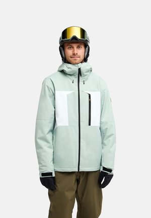 Homme portant une veste de ski vert clair et blanche, un pantalon marron, des gants noirs et des lunettes de ski teintées or sur le casque, debout devant un fond blanc.