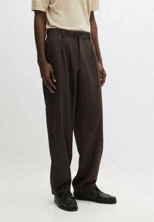 Mężczyzna w ciemnobrązowych szerokich spodniach, czarnych sznurowanych butach i beżowej koszuli z krótkim rękawem stoi na jednolitym tle.