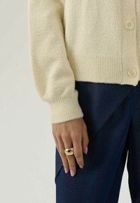 Mano con unghie rosa che indossa un anello d'oro lucido, manica di un maglione di lana color crema e pantaloni blu scuro visibili contro uno sfondo neutro.