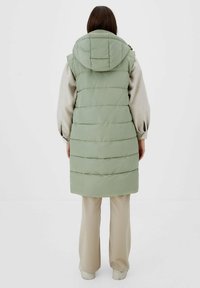 Lange donzige bodywarmer in lichtgroen met een capuchon, voorzien van horizontale stiksels. Gecombineerd met beige broek, het materiaal heeft een gladde textuur.