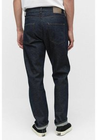 Mörkblå denimjeans med rak benpassform, featuring två bakfickor och en läderetikett på midjan. Upprullade fållar och kontrasterande sömmar synliga.