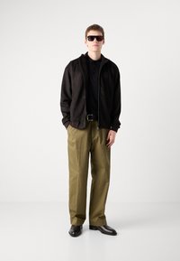 Čierna bunda na zips cez čierny polokošeľu, khaki nohavice a čierne topánky; model má na sebe hnedé, ostré slnečné okuliare. Hladké textúry látok.