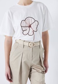 T-shirt blanc avec un grand imprimé de fleur abstraite, rentré dans un pantalon plissé taille haute beige avec une ceinture assortie.