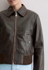 Bruine leren bomberjack met ritssluiting, twee voorzakken, geribde zoom en kraagdetails die een gladde textuur en strakke lijnen benadrukken.