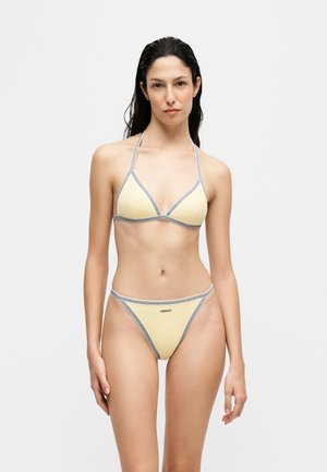 Femme aux longs cheveux foncés portant un string bikini jaune pâle avec des bordures grises et le logo Versace sur le bas, se tenant devant un fond clair uni.