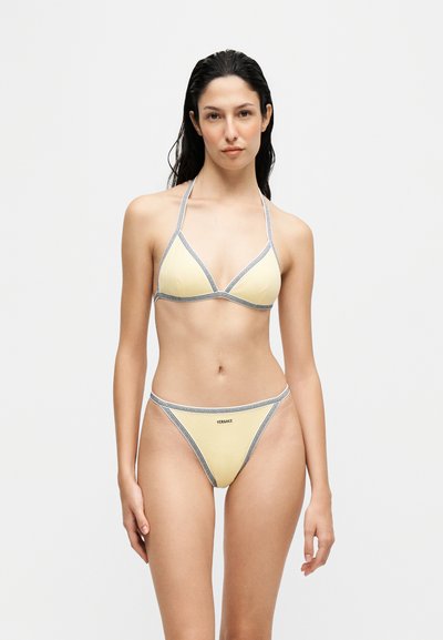 Femme aux longs cheveux foncés portant un string bikini jaune pâle avec des bordures grises et le logo Versace sur le bas, se tenant devant un fond clair uni.