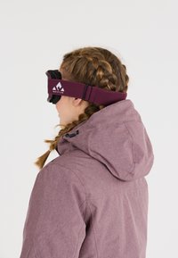 Skibril met een zwart frame en een kastanjebruine verstelbare band met een wit logo, gedragen door een persoon in een paarse jas met capuchon.