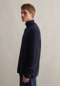 Granatowy sweter z dzianiny z wysokim kołnierzem i półzamkiem, z teksturalnym, waflowym wzorem oraz ściągaczami przy mankietach.