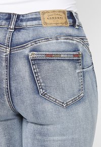 Verblasste blaue Denim-Jeans mit einem Lederpatch, dekorativen Nähten und einer kleinen, gemusterten Gesäßtasche mit silbernen Nieten.