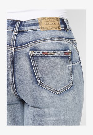 Išblukę mėlyni džinsai su odiniu užtrauktuku, dekoratyvine siuvinėjimu ir maža, raštuota nugara kišene su sidabriniais varžtais.