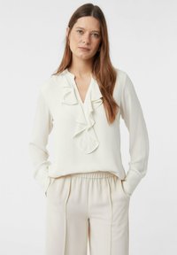 Biała, jedwabna bluzka z dekoltem w serek i falbanami wzdłuż przodu, zestawiona z beżowymi spodniami z elastycznym pasem.