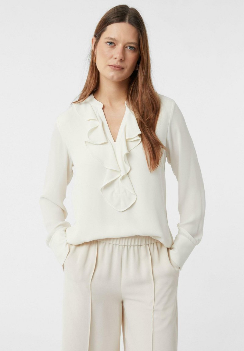 Biała, jedwabna bluzka z dekoltem w serek i falbanami wzdłuż przodu, zestawiona z beżowymi spodniami z elastycznym pasem.