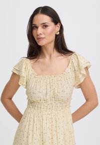Femme aux longs cheveux foncés portant de petites boucles d'oreilles en or et une robe jaune pâle à motifs floraux avec des manches à volants et un décolleté carré.