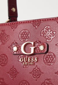 Burgundowa torebka wzorzysta z wytłoczonymi wzorami, złotymi akcentami i paskiem. Zawiera logo „G” oraz „GUESS EST 1981” na metce.