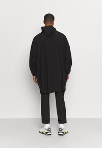 Schwarzer kapuzen Poncho mit lockerer Passform, breiten Ärmeln und Druckknöpfen an den Seiten, getragen über schwarzen Hosen und Sneakers.