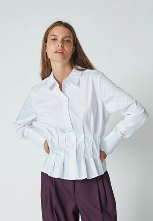 White cotton button-up shirt featuring a pleated hem and long sleeves, paired with formal purple trousers. Smooth texture and structured design.