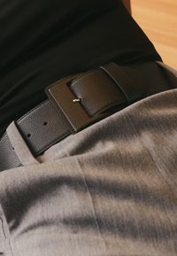 Juodas odinis diržas su stačiakampiu sagtimi, dėvimas virš pilkų audinių kelnių, pasižymintis lygiu paviršiumi ir subtiliu blizgesiu.