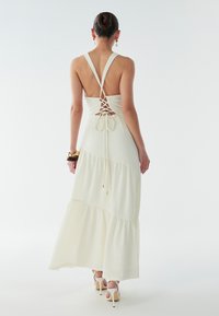 Lange witte jurk met een gestructureerde stof, open rug met kruislingse bandjes en vallende ruches aan de zoom.