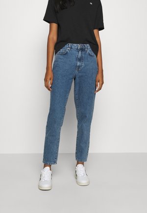 Persoon draagt een blauwe jeans met hoge taille, een zwart T-shirt met korte mouwen en witte sneakers, staand tegen een lichte, effen achtergrond.