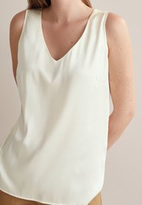 Top senza maniche a scollo a V in tessuto crema chiaro. Texture liscia con una vestibilità rilassata e un piccolo dettaglio tasca sul lato sinistro.